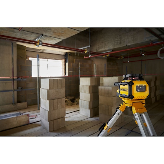 A DEWALT rotary laser level on a tripod is set up in a construction site with unfinished walls and concrete blocks. The laser emits a horizontal red line across the room to assist with accurate leveling and alignment work.