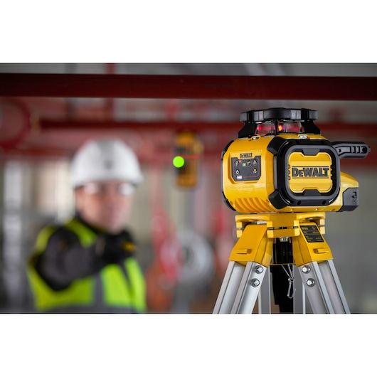A DEWALT rotary laser level (SKU: DCLE15211R) is mounted on a tripod in a construction environment. The laser device is yellow and black with control buttons visible. In the background, a blurred person wearing a safety vest and helmet is seen, along with industrial pipes.