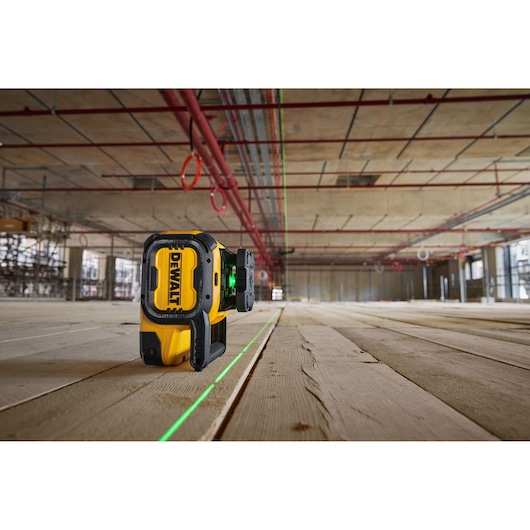 A DEWALT green laser level device placed on a wooden floor in a large, unfinished construction site, projecting a visible green laser line across the floor.