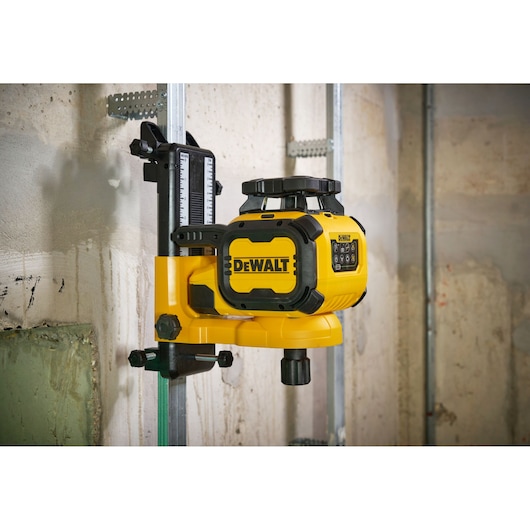 A yellow DEWALT rotary laser level mounted on a metal bracket, attached to a vertical metal rail, in an industrial or construction setting with concrete walls.