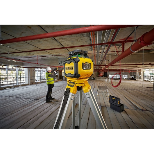 A DEWALT laser level (model DCLE15221G) is mounted on a tripod inside a construction site. A worker wearing a hard hat and safety vest is seen in the background measuring or inspecting the area. There are pipes and construction materials visible throughout the open, unfinished indoor space.