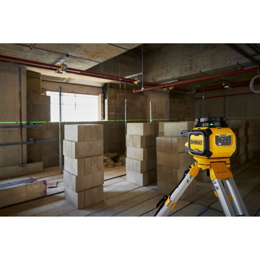 A DEWALT laser level device is positioned on a tripod in a construction site, projecting a green horizontal line across the unfinished room with concrete blocks and exposed pipes.