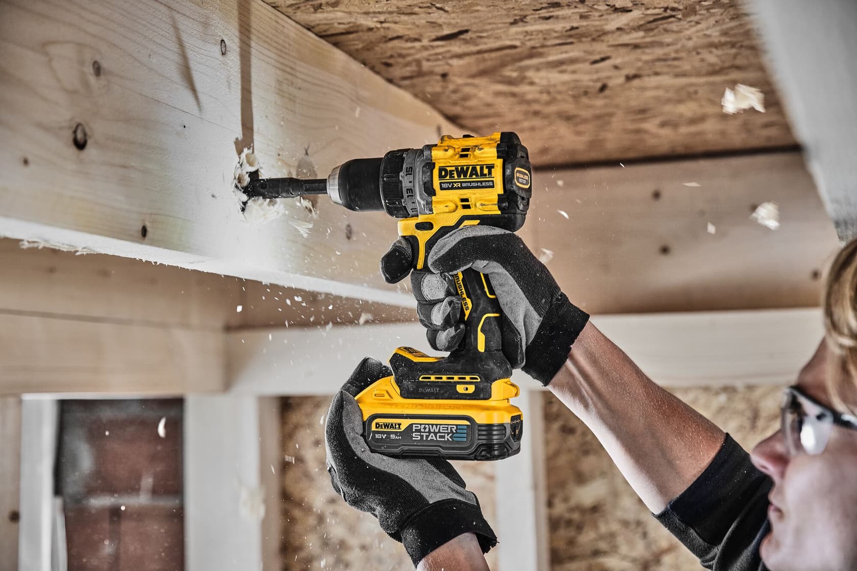 Una persona usando gafas de seguridad y guantes utiliza un taladro inalámbrico DEWALT para perforar un agujero en una viga de madera, mientras los restos de madera vuelan durante el proceso.
