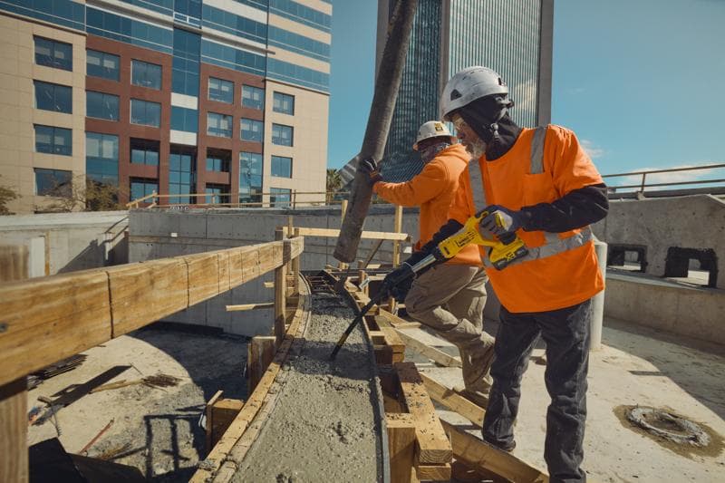 Dos trabajadores de construcción con cascos de seguridad y camisetas naranjas de alta visibilidad trabajan en una obra. Uno usa una herramienta vibradora de concreto DEWALT sobre concreto recién vertido, mientras el otro dirige una manguera de concreto. Edificios de oficinas modernos se ven al fondo.