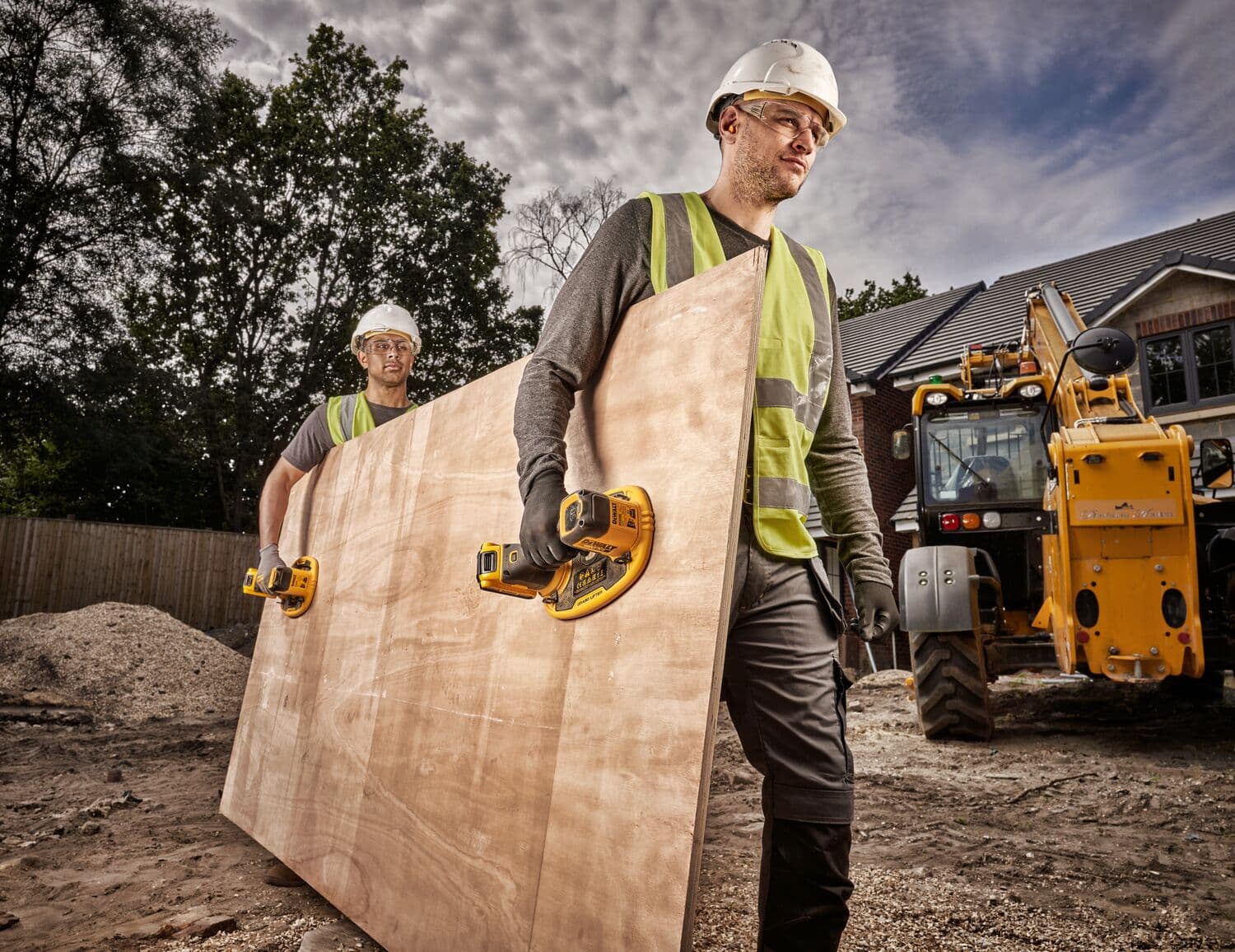 Dos trabajadores de la construcción con chalecos reflectantes y cascos utilizan elevadores de ventosa inalámbricos DEWALT para transportar una gran placa de madera contrachapada en una obra. Se ve un vehículo de construcción amarillo y una casa al fondo.