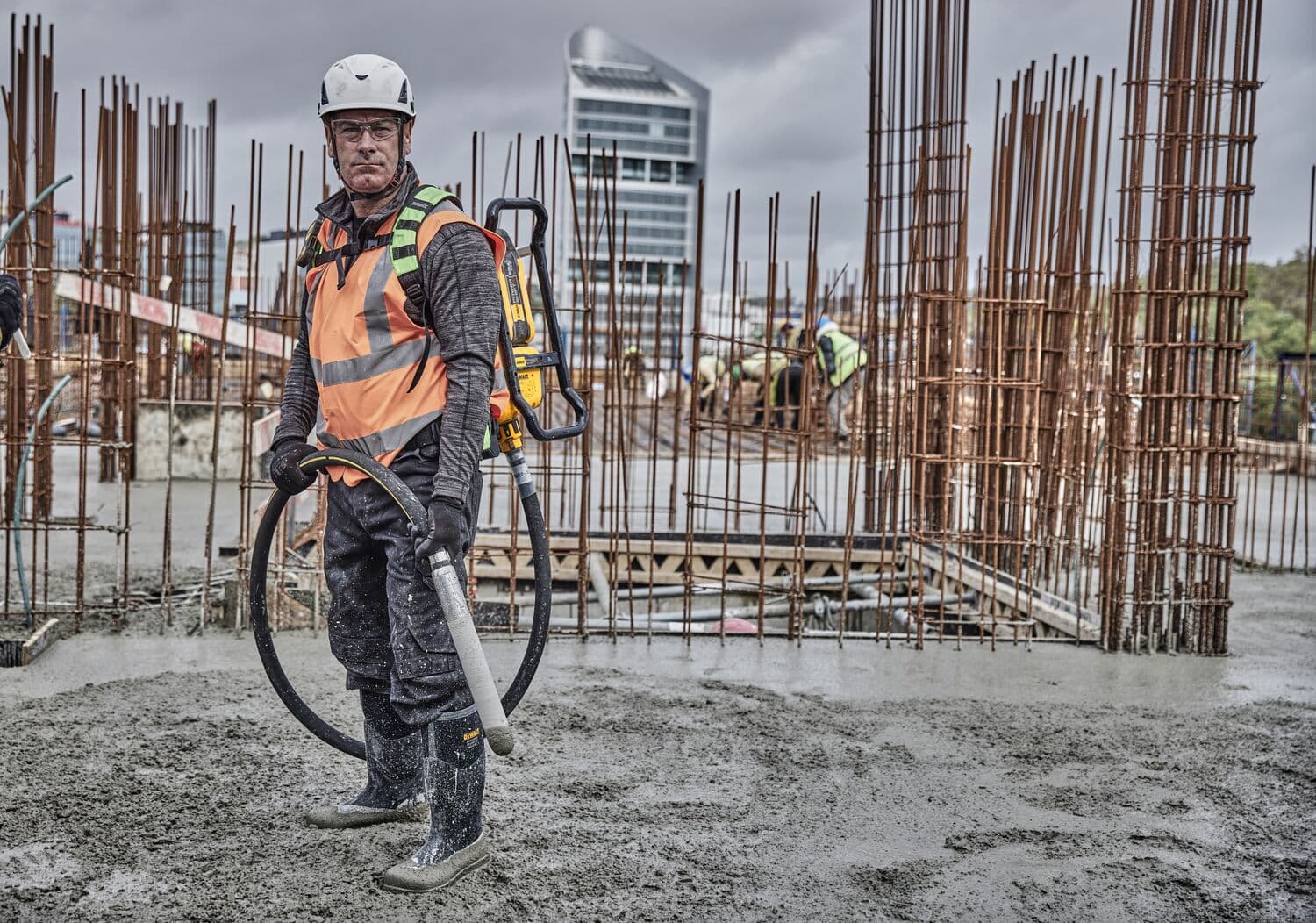 A construction worker wearing safety gear and an orange high-visibility vest operates a DEWALT concrete vibrator backpack (SKU: DCPS310N) at a building site with rebar structures and wet concrete. The background includes other workers and an urban building.