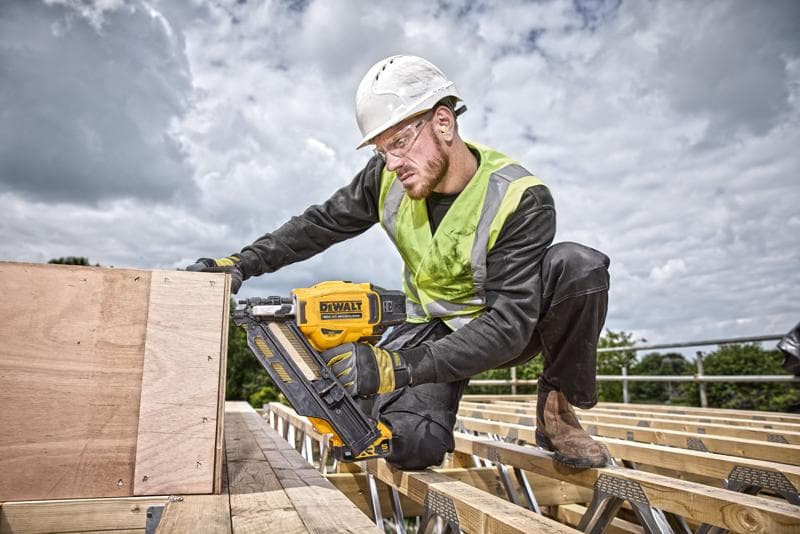 Un trabajador de la construcción con casco de seguridad y chaleco de alta visibilidad utiliza una clavadora amarilla DEWALT DCN930N para fijar tablas de madera en un sitio de construcción al aire libre. El rostro del trabajador está desenfocado para mantener la privacidad.