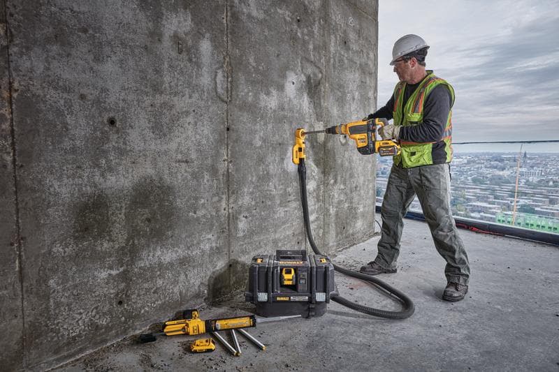 Un trabajador de la construcción con chaleco de seguridad y casco está usando un taladro inalámbrico DEWALT con accesorio de extracción de polvo para perforar una pared de hormigón. El trabajador está sobre un edificio grande con una vista urbana de fondo. Hay varias herramientas DEWALT y una caja de herramientas colocadas cerca en el suelo.