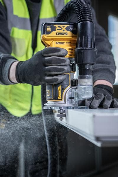 Una persona con guantes de seguridad y chaleco de alta visibilidad utiliza una fresadora inalámbrica DEWALT con extracción de polvo, trabajando en el borde de una pieza de madera u otro material plano.
