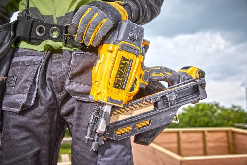 Primer plano de una persona con guantes de trabajo sosteniendo una clavadora DEWALT DCN930N en un sitio de construcción, con una estructura de madera al fondo.