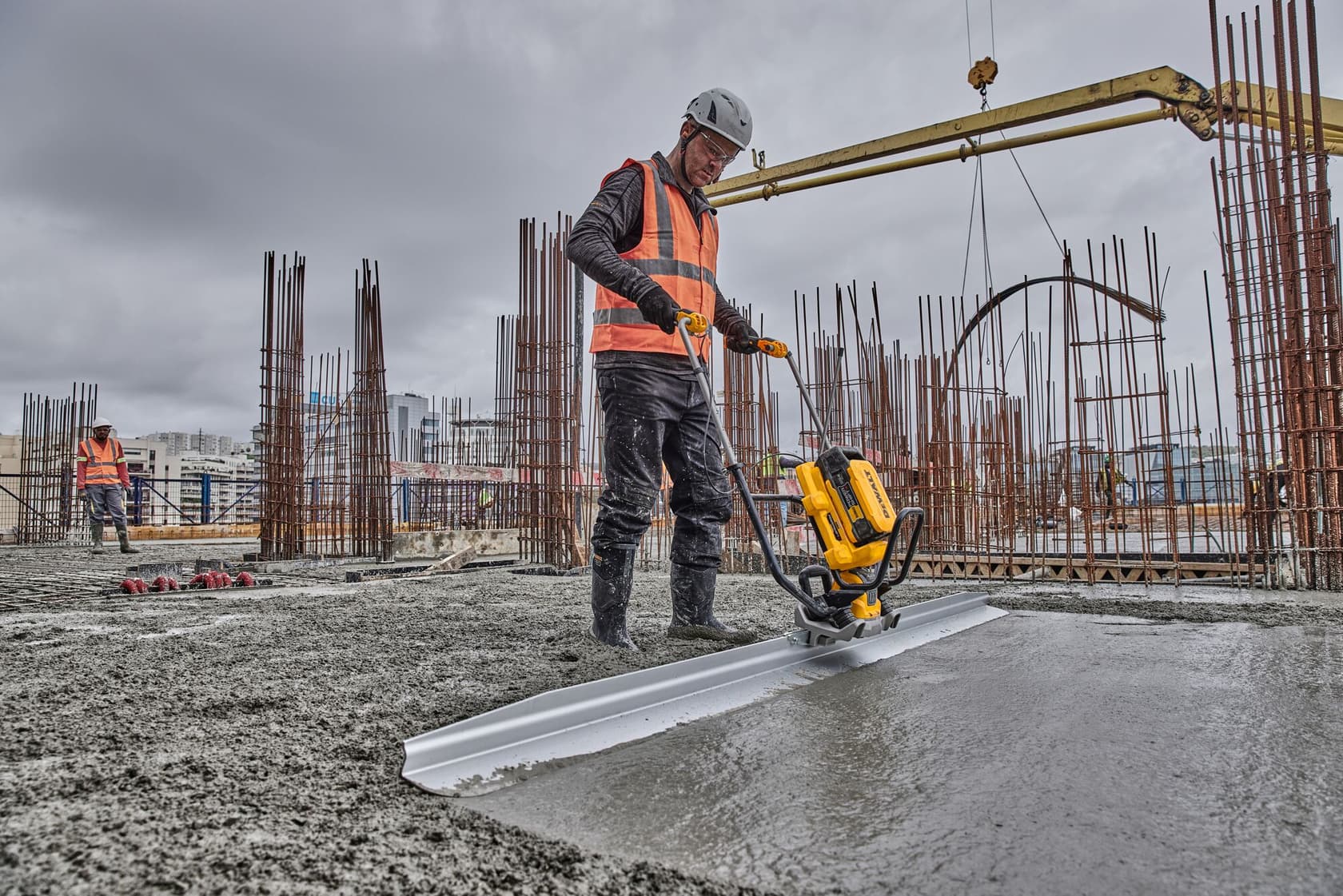 A construction worker wearing a helmet and safety vest is operating a yellow DEWALT concrete screed machine to level wet concrete on a building site with structural steel rods in the background. The worker's face is not identifiable.