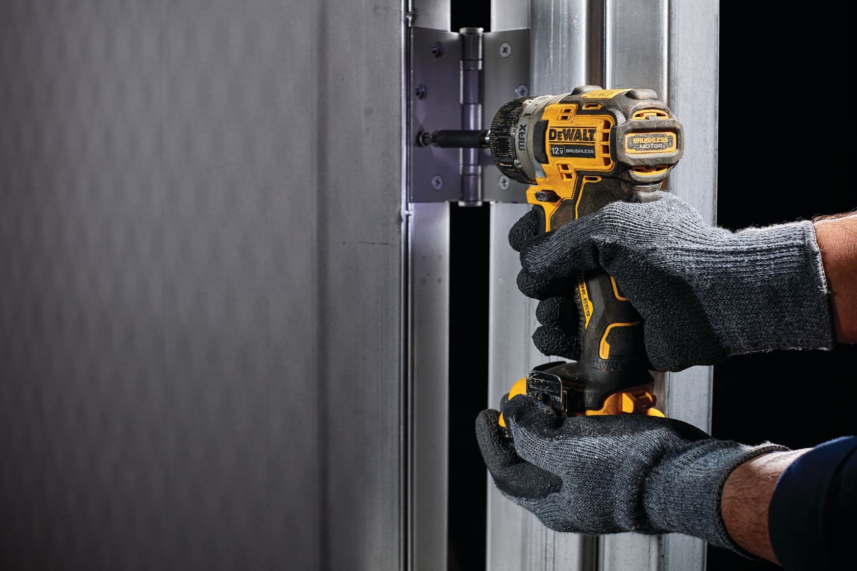 Una persona con guantes grises está usando un taladro inalámbrico amarillo DEWALT para instalar o apretar tornillos en una bisagra de metal conectada a un marco de puerta metálico.