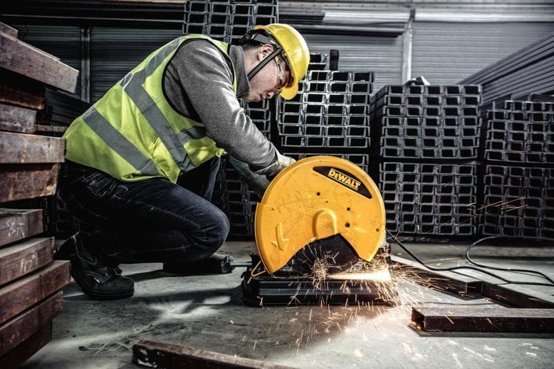 Un trabajador con chaleco de seguridad y casco utiliza una herramienta eléctrica DEWALT D28730 para cortar metal, generando chispas en un entorno industrial con vigas de metal apiladas.