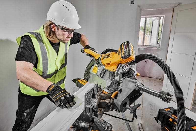 Una persona con chaleco de seguridad, guantes y casco está utilizando una sierra de inglete inalámbrica DEWALT DCS781X2 para cortar una moldura dentro de una habitación en construcción. La sierra está conectada a un sistema de extracción de polvo y se pueden ver baterías de herramientas DEWALT cerca.