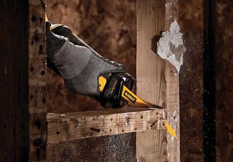 Una mano enguantada usando una hoja de sierra recíproca DEWALT para cortar un marco de madera, con aserrín y escombros volando alrededor.