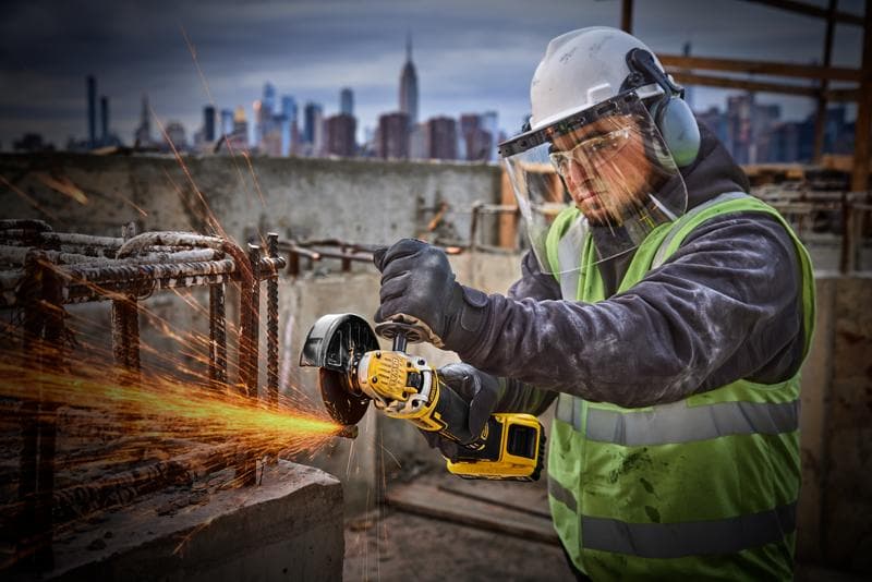 Una persona con equipo de protección, incluyendo casco, protector facial, guantes y chaleco reflectante, usa una amoladora angular inalámbrica amarilla DEWALT (SKU: DCG405P2) para cortar barras de metal en un sitio de construcción. Salen chispas de la zona de corte. El horizonte de la ciudad se ve en el fondo.