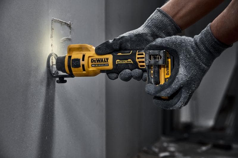 Una persona con guantes grises está usando una herramienta rotativa inalámbrica DEWALT para cortar un agujero cuadrado en un panel de yeso.