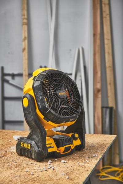 Un ventilador portátil inalámbrico DEWALT (modelo DCE512B) está colocado sobre un banco de trabajo de madera en un taller con materiales de construcción al fondo.