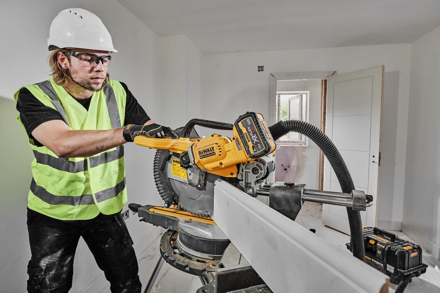 Una persona con casco de seguridad y chaleco reflectante opera una sierra ingletadora inalámbrica DEWALT DCS781N, cortando un trozo de madera en un entorno de construcción o renovación.