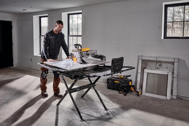 Una persona está al lado de una sierra para azulejos DEWALT D36000S montada en su soporte en una habitación en proceso de construcción. La sierra está sobre un soporte plegable negro y hay herramientas y una bolsa de herramientas DEWALT en el suelo cerca. La habitación tiene paredes sin terminar, ventanas y materiales de construcción apoyados contra la pared.