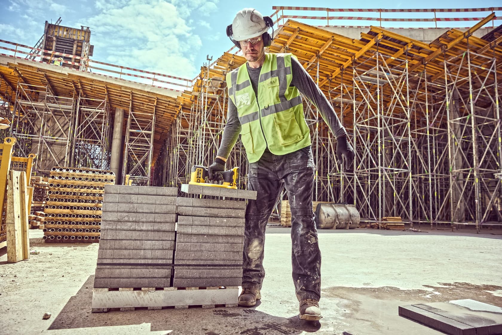A construction worker wearing a safety vest, hard hat, and ear protection is using a DEWALT DCE590 tool to lift a concrete slab from a stack at a construction site. Scaffolding and building materials are visible in the background.