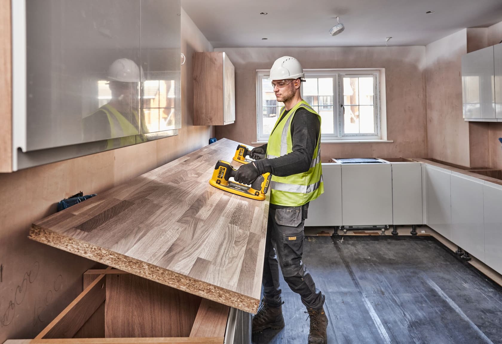 Un trabajador de la construcción con casco blanco y chaleco de seguridad amarillo utiliza una herramienta amarilla DEWALT para levantar una encimera de madera en una cocina moderna en renovación.