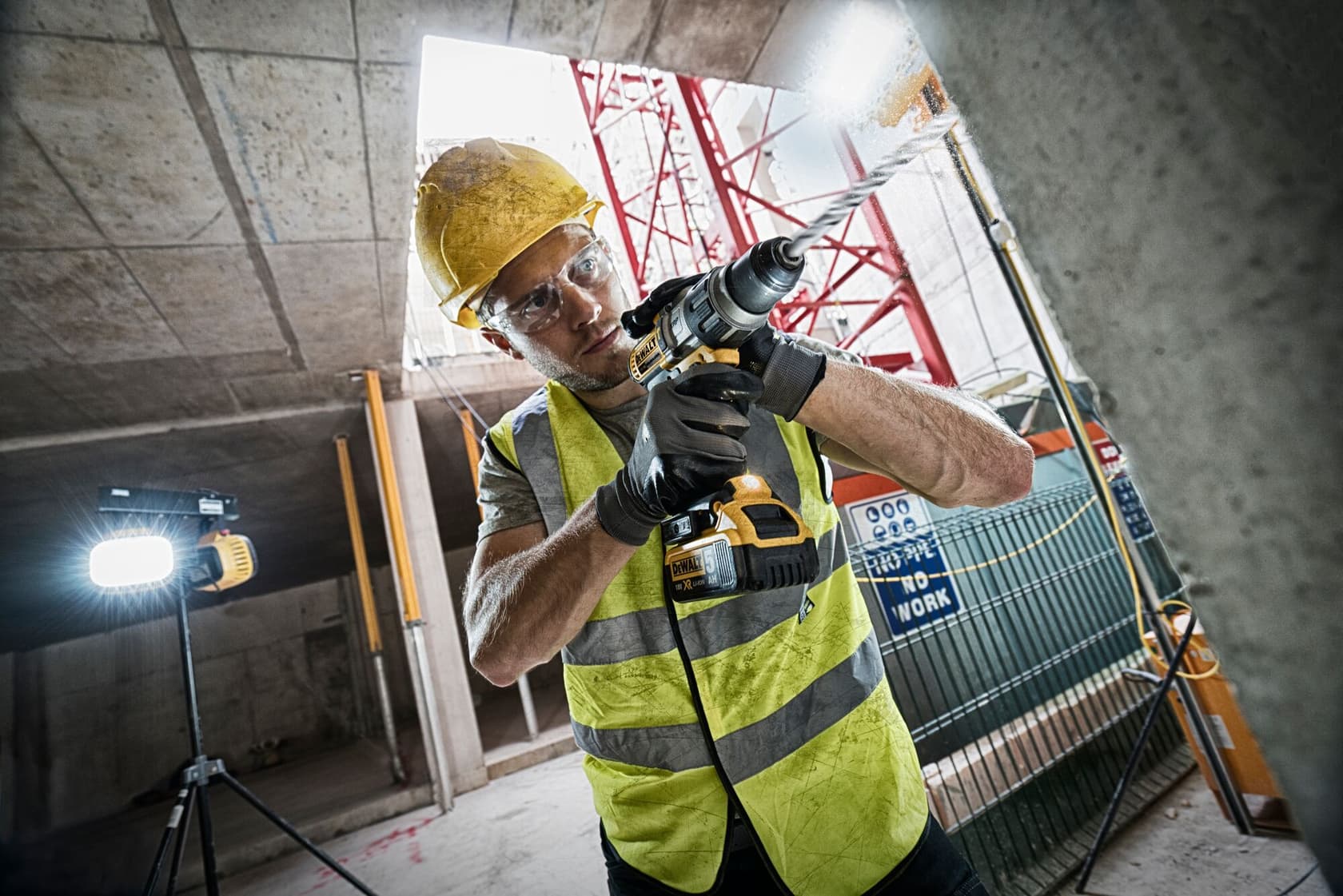 Un trabajador de la construcción con casco amarillo, chaleco reflectante y guantes negros utiliza un taladro inalámbrico en una obra. Hay equipo de construcción y una luz en el fondo.