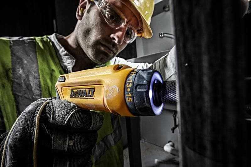 Una persona con chaleco de seguridad reflectante y guantes utiliza un destornillador inalámbrico amarillo DEWALT (modelo DCF680G2) para trabajos de mantenimiento en interiores. El enfoque está en la herramienta y la mano enguantada del usuario. El rostro de la persona está difuminado para proteger su privacidad.