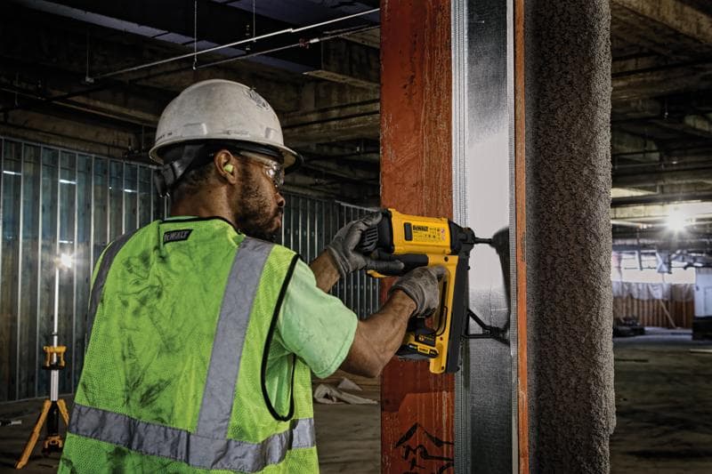 Un trabajador de construcción con casco, protección auditiva, guantes y chaleco reflectante utiliza una clavadora DEWALT para fijar un perfil metálico a una viga de acero en una obra interior.