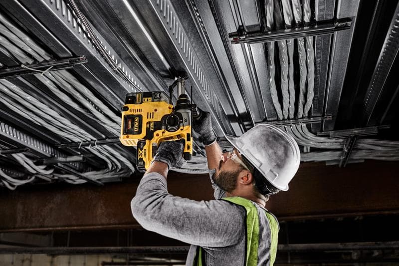 Una persona con casco, gafas de seguridad, guantes y chaleco de seguridad utiliza un taladro rotatorio inalámbrico amarillo DEWALT para perforar un techo de metal con cables eléctricos.