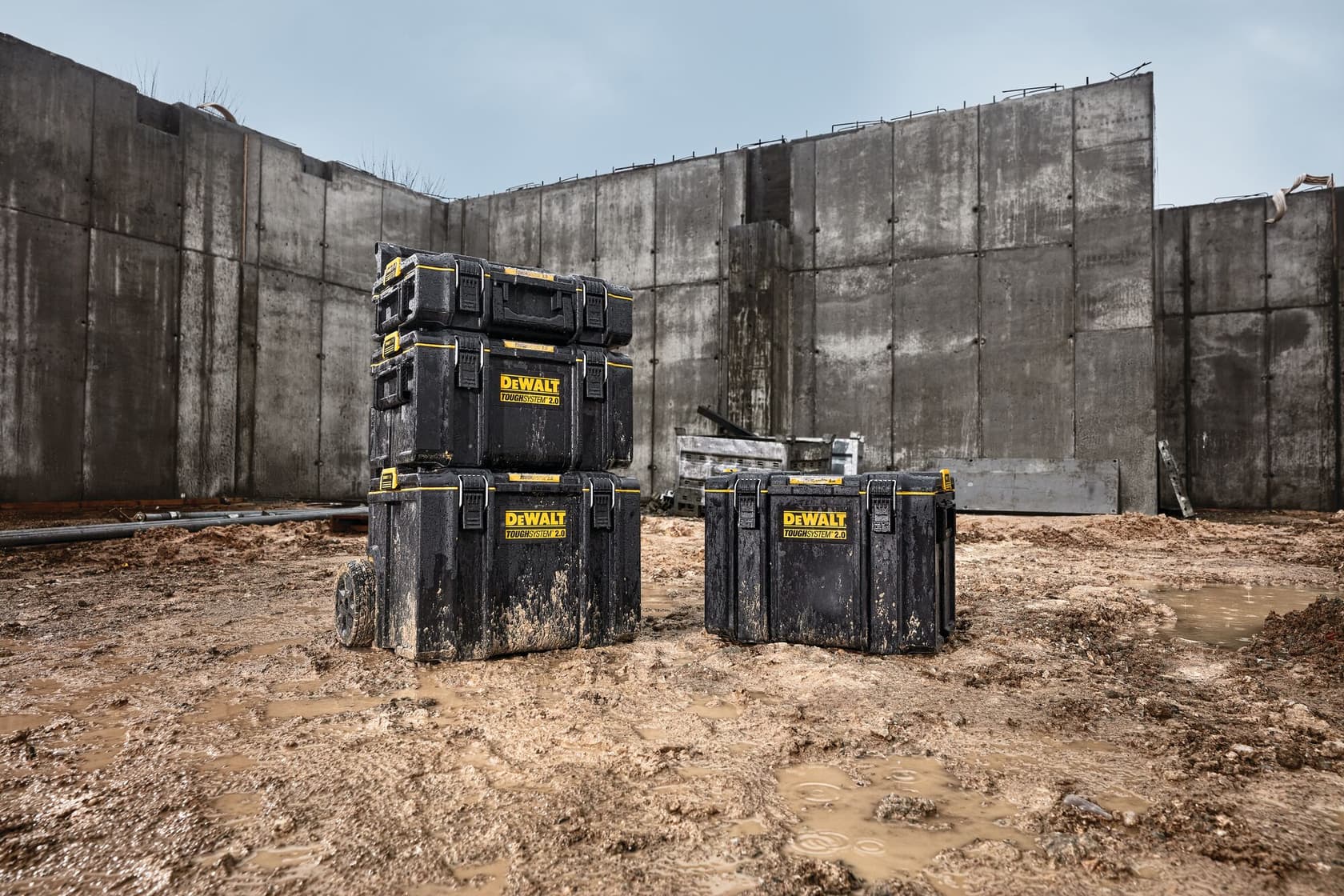 Tres cajas de almacenamiento DEWALT ToughSystem negras están colocadas en un sitio de construcción embarrado, dos apiladas a la izquierda y una a la derecha, con altos muros de concreto en el fondo.
