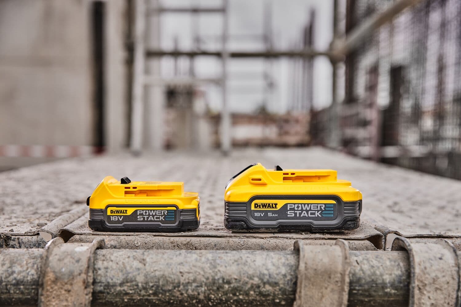 Dos baterías DEWALT POWERSTACK de 18V se muestran una al lado de la otra en un sitio de construcción, con un diseño amarillo y negro. Las baterías están colocadas sobre una superficie metálica con un fondo industrial desenfocado.