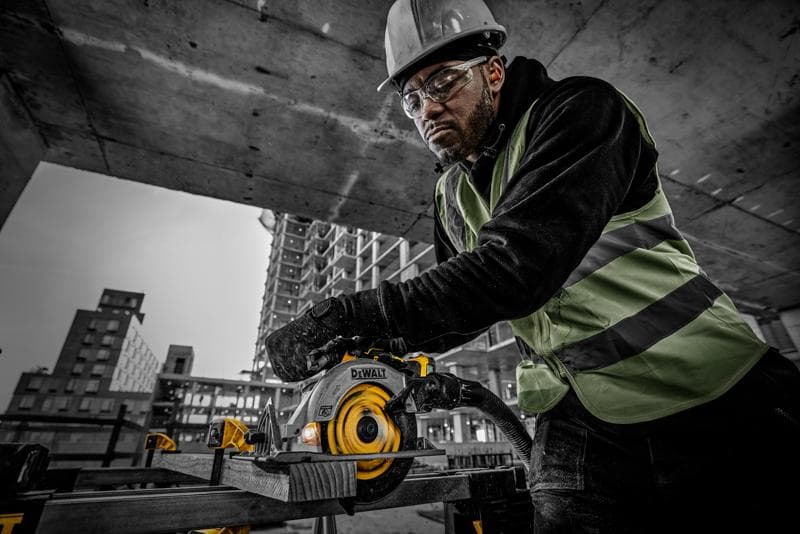 Un trabajador de la construcción con chaleco de seguridad y casco opera una sierra circular DEWALT (modelo DCS565NT) para cortar una tabla de madera en un sitio de construcción. Al fondo se observan estructuras de concreto y edificios en construcción.