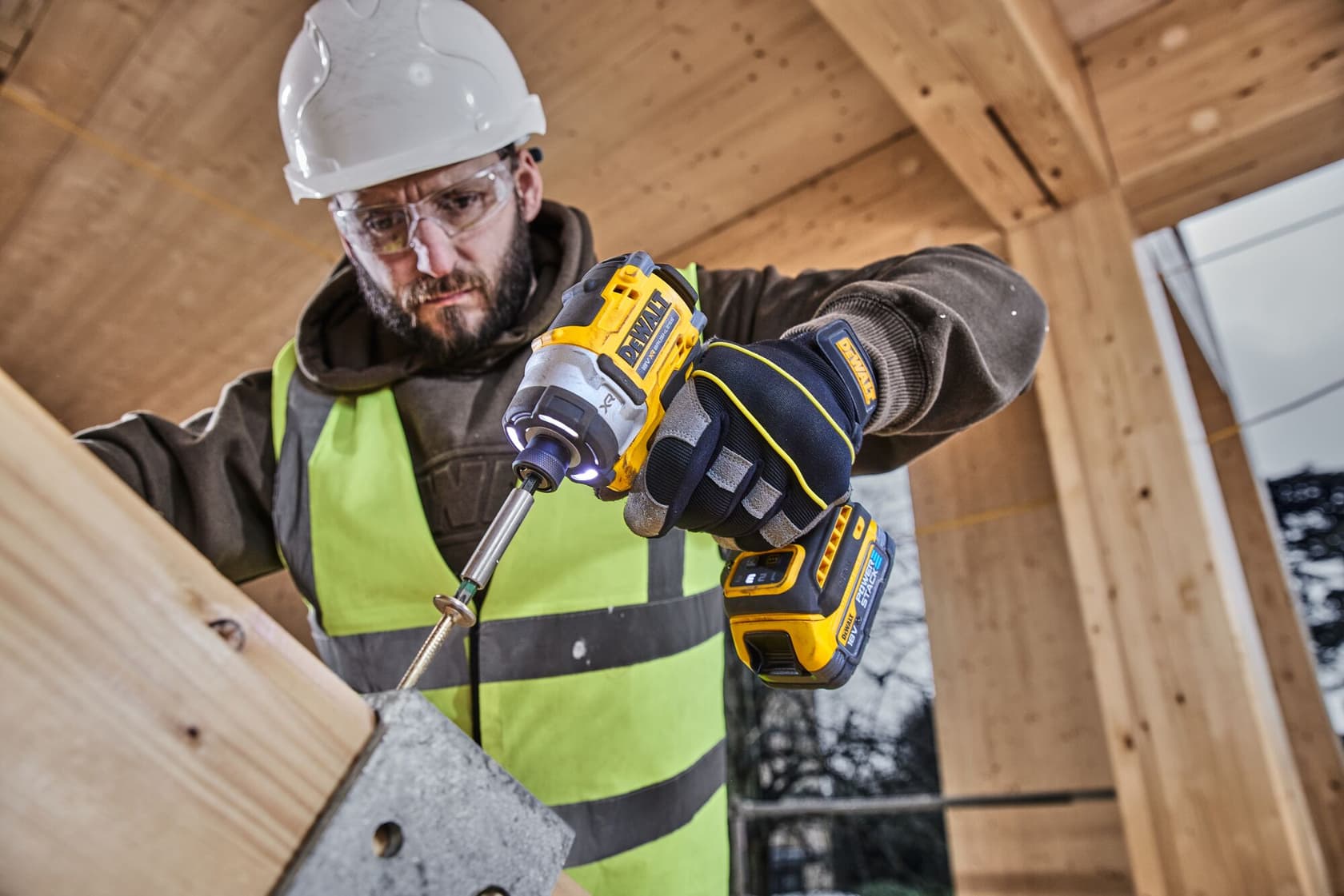 A person wearing a safety vest, gloves and hard hat is using a DEWALT DCF860NT cordless impact driver to drive a screw into a piece of wood, inside a wooden structure.