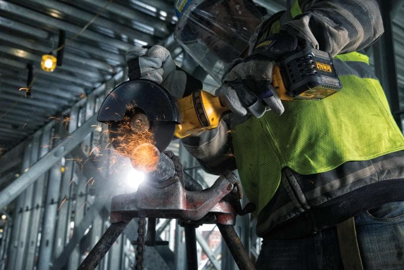 Primer plano de una persona con guantes protectores y chaleco de seguridad usando una amoladora angular inalámbrica DEWALT para cortar metal, con chispas volando, en un sitio de construcción industrial.
