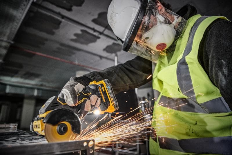 Un trabajador con equipo de protección, incluyendo casco, pantalla facial, guantes y chaleco de alta visibilidad, utiliza una amoladora angular inalámbrica DEWALT (modelo DCS438N) para cortar metal, generando chispas.