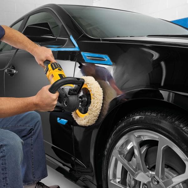 Una persona utiliza una pulidora DEWALT DWP849X sobre la superficie negra de un automóvil, concentrándose en el área sobre el neumático delantero. La pulidora tiene diseño amarillo y negro y una almohadilla de lana grande. Se ha colocado cinta azul en algunas partes del automóvil para protegerlas.