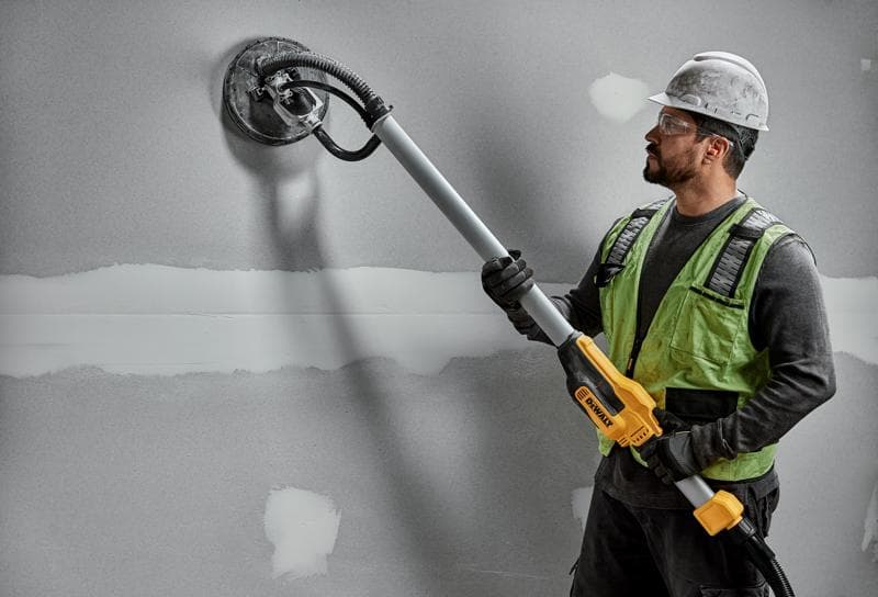 Un trabajador de la construcción con casco y chaleco de seguridad utiliza una lijadora de paneles de yeso DEWALT DWE7800 con mango telescópico para lijar una pared gris.