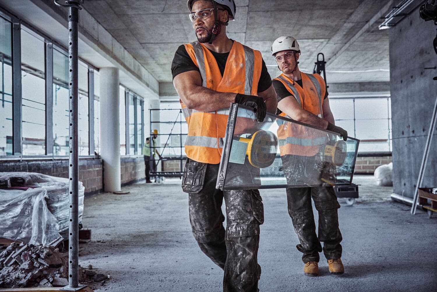 Dos trabajadores de la construcción con chalecos de seguridad naranjas y cascos llevan una gran lámina de vidrio con una herramienta de ventosa DEWALT dentro de un edificio en construcción.