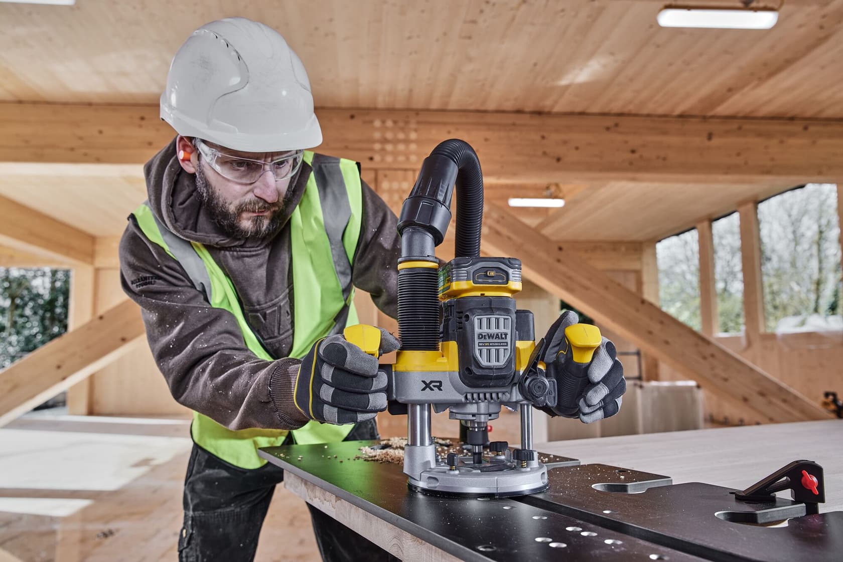 A worker wearing a safety helmet and high-visibility vest operates a DEWALT DCW620NT cordless router on a flat surface, inside a wooden construction site. The router is attached to a dust extraction hose, and wood shavings are visible on the work surface.