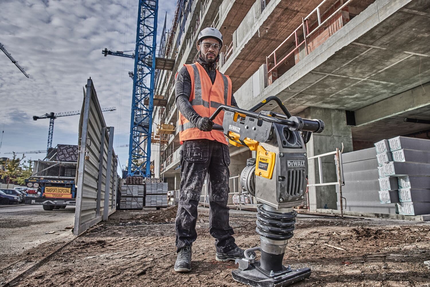 A construction worker wearing a safety vest and helmet operates a DEWALT DCPS660N power tool on a construction site, with a partially constructed building and cranes visible in the background.