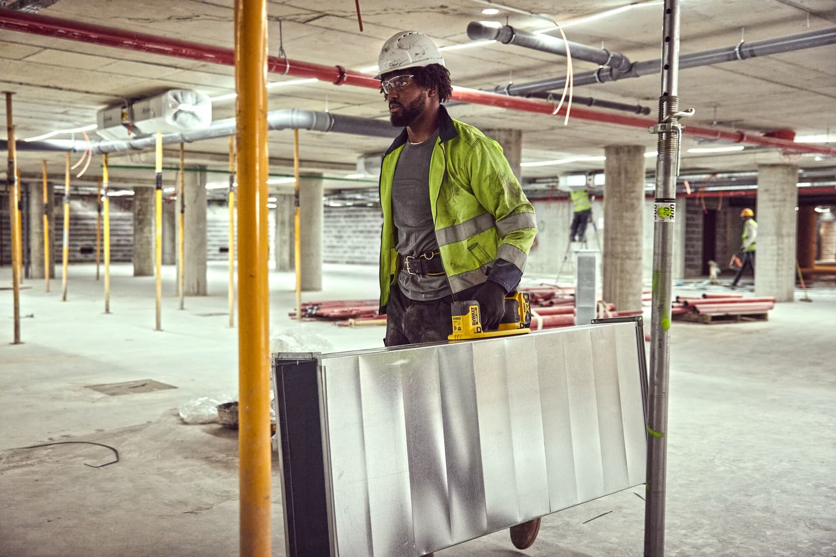 A person in a high-visibility jacket and hard hat is carrying a large metal panel using a DEWALT tool in a construction site with concrete pillars and pipes. The person’s face is blurred.