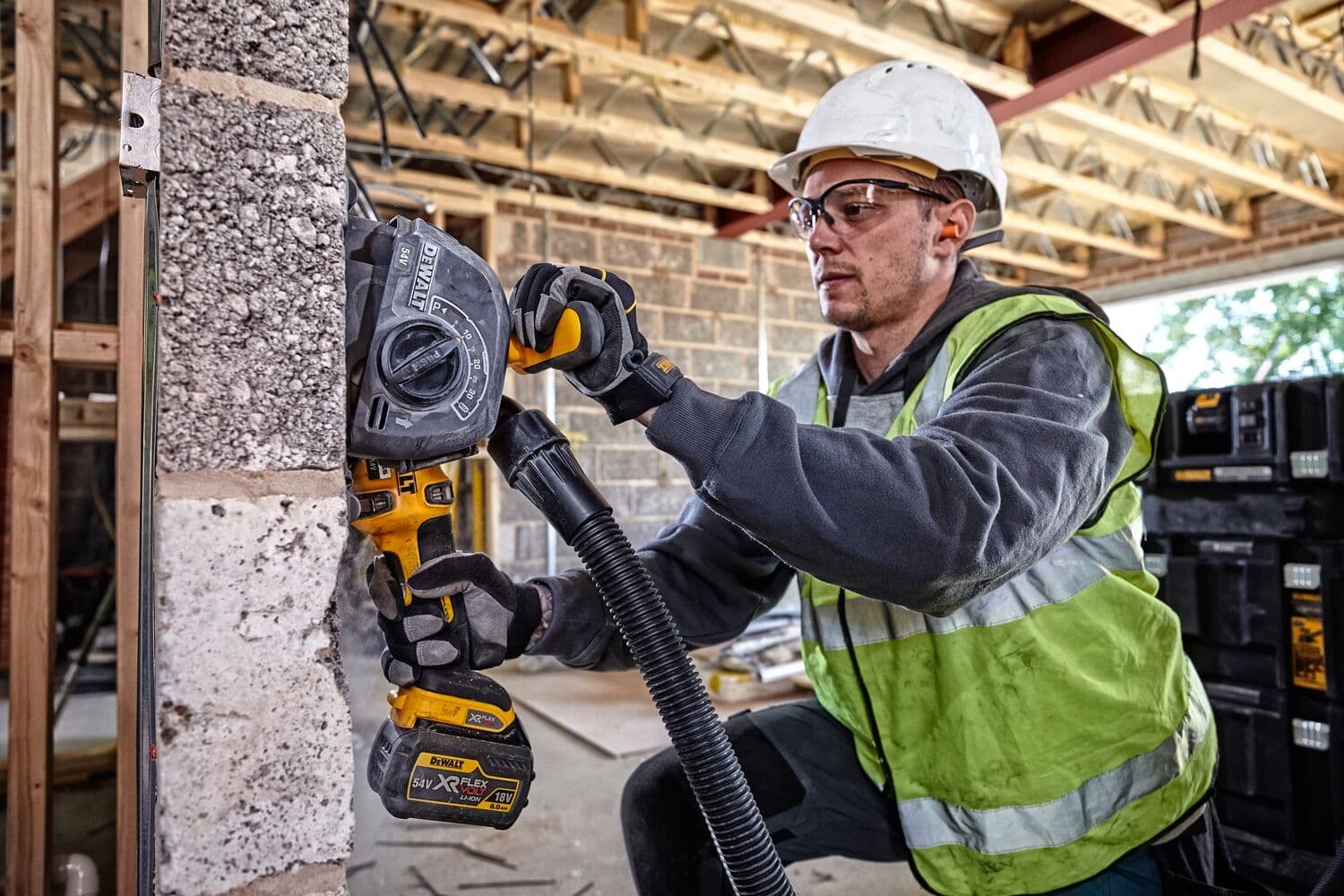 Persona con casco de seguridad y chaleco de alta visibilidad utilizando una herramienta DEWALT mural inalámbrica en una pared de bloques de hormigón en un sitio de construcción. La herramienta tiene una manguera de extracción de polvo. Cajas de almacenamiento DEWALT visibles al fondo.