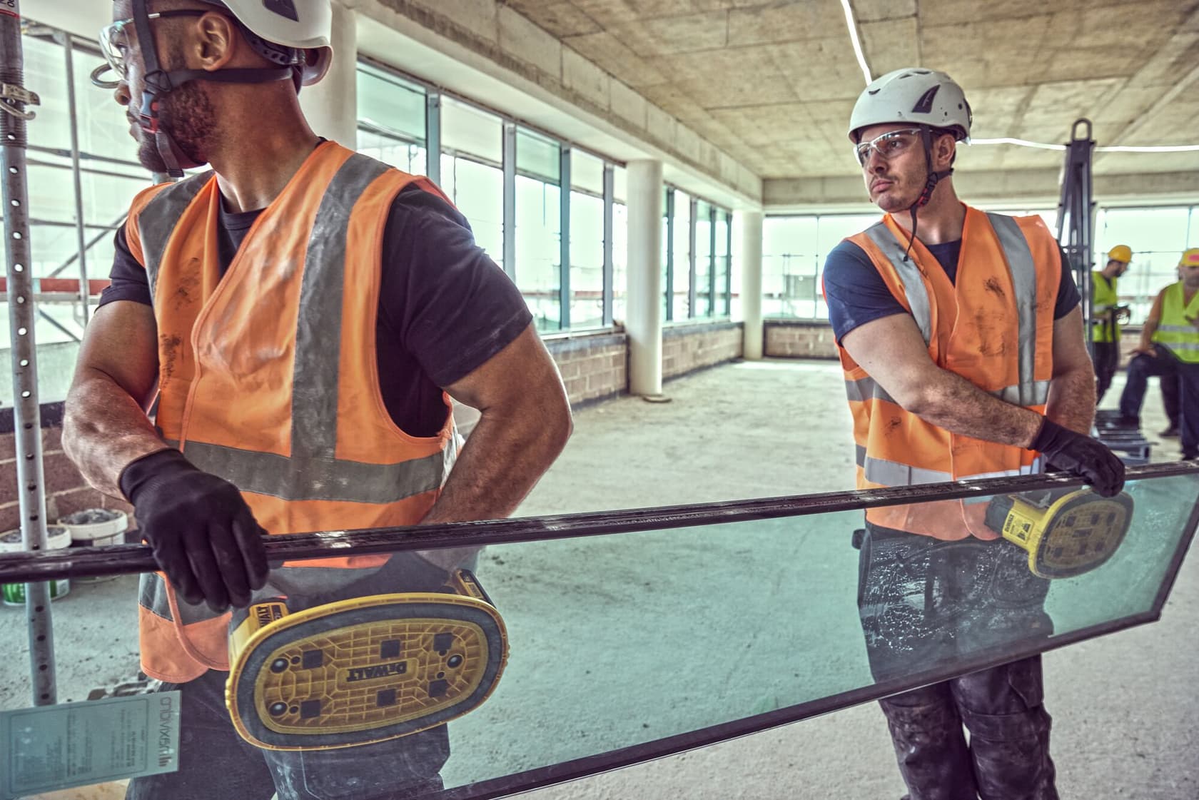 Two construction workers wearing safety helmets, gloves, and orange reflective vests are using DEWALT DCE590 tools to lift and carry a large glass panel at an indoor construction site. Other workers and construction materials are visible in the background.