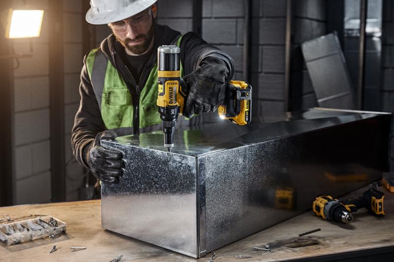 Una persona con equipo de seguridad utiliza una herramienta eléctrica inalámbrica DEWALT DCF403NT para trabajar en una caja de metal en un taller industrial. Se ven varios tornillos y herramientas sobre la mesa de trabajo.