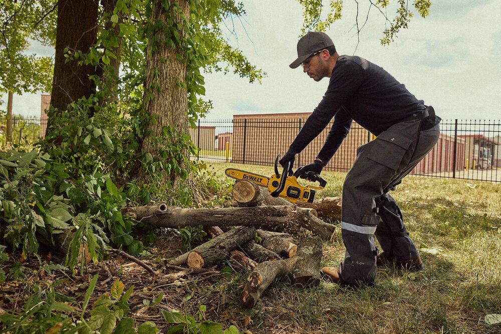 Una persona con ropa protectora y guantes está utilizando una motosierra inalámbrica amarilla DEWALT para cortar ramas o troncos al aire libre cerca de un árbol.