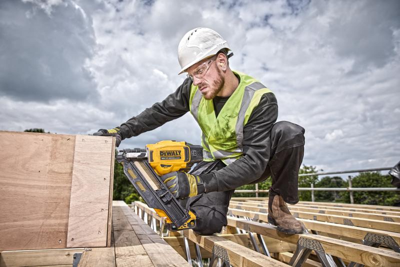 Un trabajador de la construcción con casco de seguridad y chaleco de alta visibilidad utiliza una clavadora amarilla DEWALT DCN930N para fijar tablas de madera en un sitio de construcción al aire libre. El rostro del trabajador está desenfocado para mantener la privacidad.