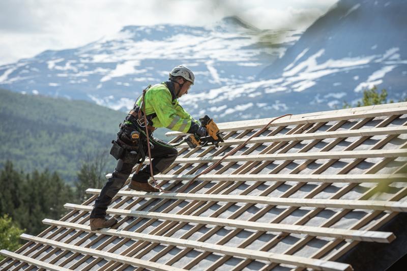 Un trabajador de la construcción con equipo de seguridad utiliza una clavadora inalámbrica DEWALT para fijar listones de madera en un tejado inclinado, con un paisaje montañoso de fondo.
