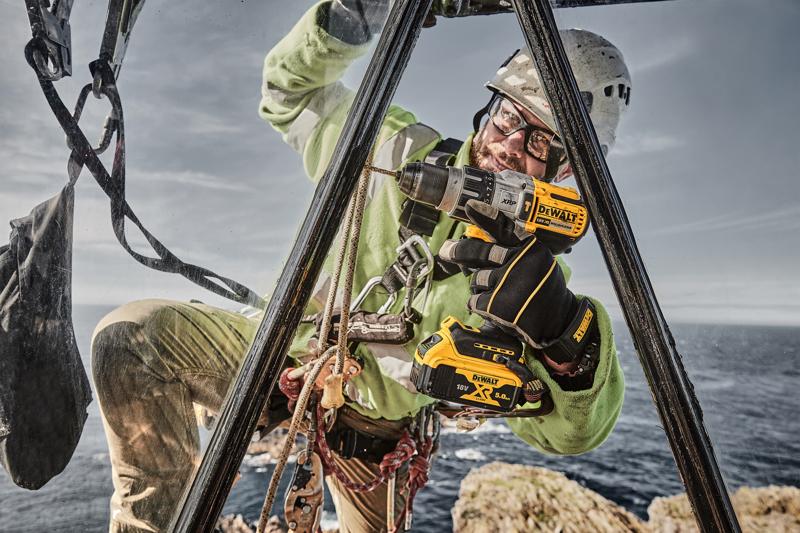 Un trabajador equipado con equipo de seguridad utiliza un taladro inalámbrico DEWALT DCD996P2 al aire libre, situado en una estructura metálica cerca de un acantilado, con el mar de fondo.