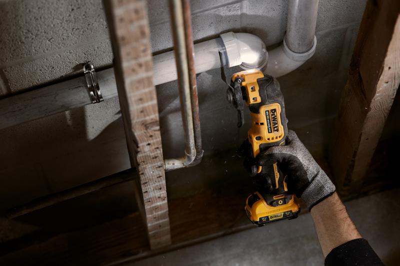 Una persona con guante utiliza una herramienta multifunción oscilante inalámbrica DEWALT para cortar una tubería de PVC blanca cerca de una pared con tubos y vigas expuestas.